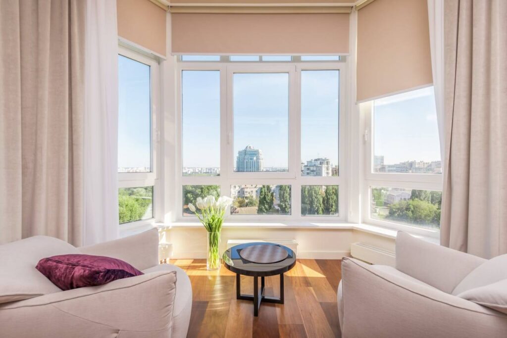 Bright living room with large windows, city view, cozy chairs, and a small table with flowers.