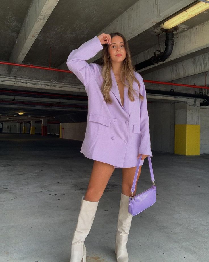 Woman in lavender blazer and white boots holding a matching purse in a parking garage. Fashionable and stylish outfit.