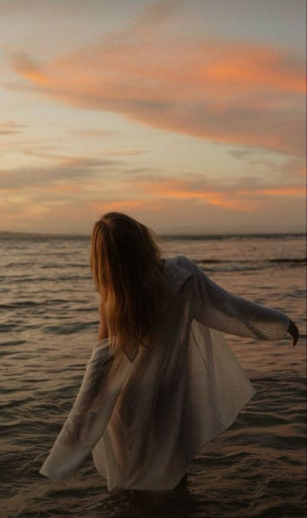 Person in white dress stands in calm ocean at sunset, with vibrant sky and flowing hair. Peaceful beach scene.