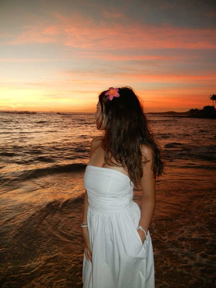 Woman in white dress on beach at sunset, with flower in hair, looking at ocean waves.