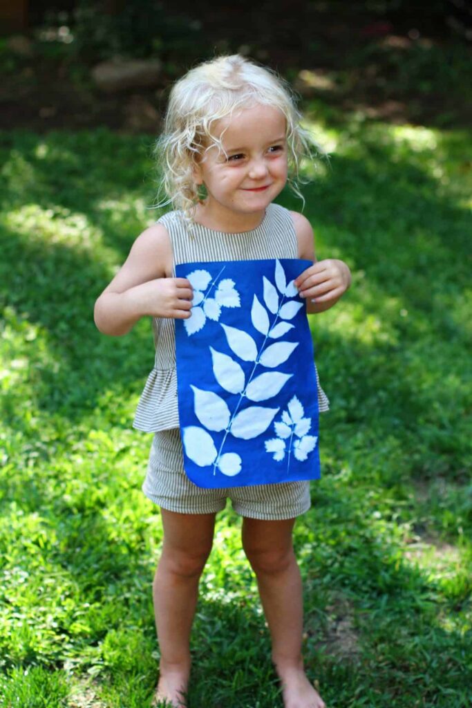 Child holding blue leaf print fabric outdoors on grass.