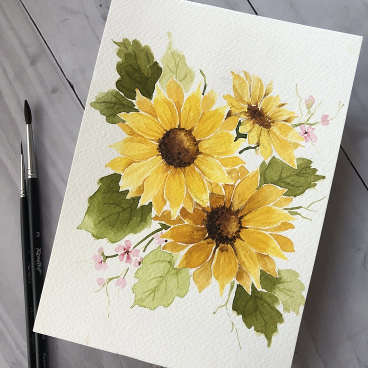 Watercolor painting of yellow sunflowers with green leaves on a white paper beside paintbrushes.