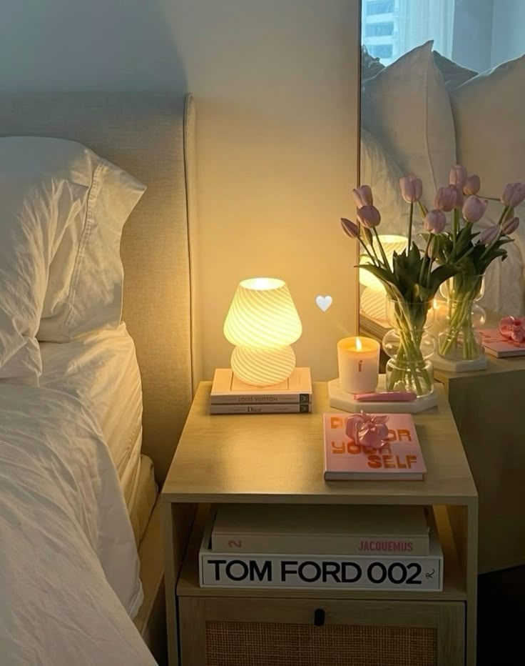 Cozy bedroom with side table lamp, pink tulips, candle, and books for a serene, stylish ambiance.