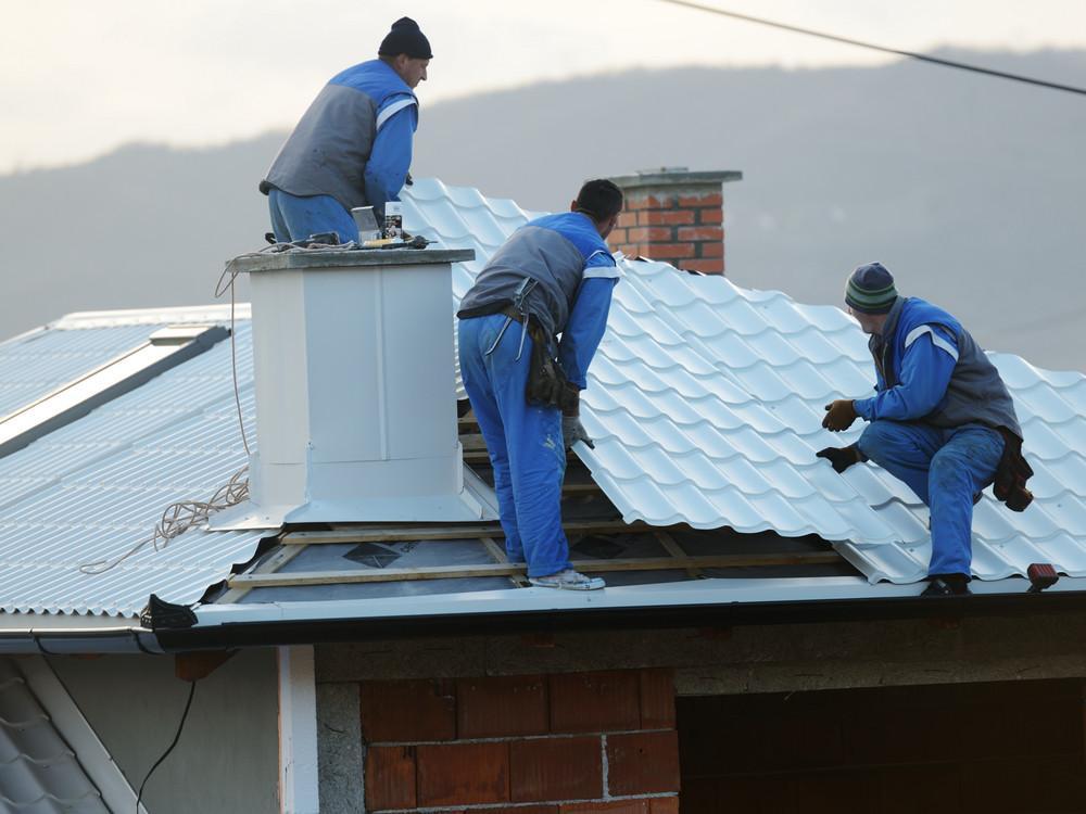 Workers installing metal roofing on a house with chimney in the background.