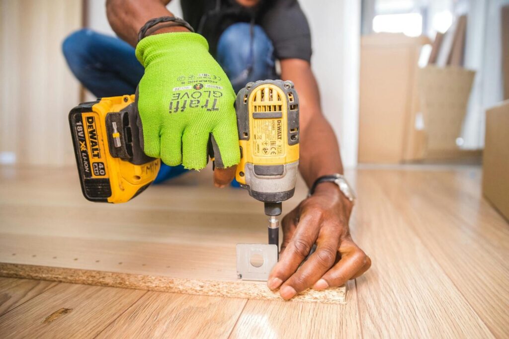 Person in green gloves using a DeWalt power drill on wooden floor assembly. DIY home improvement.