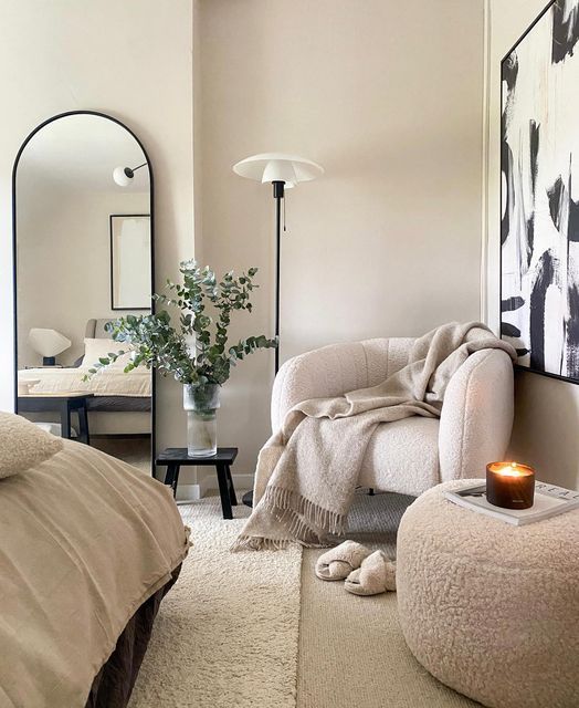 Cozy beige bedroom with plush chair, blanket, candle, and modern decor. Relaxing interior design with greenery.