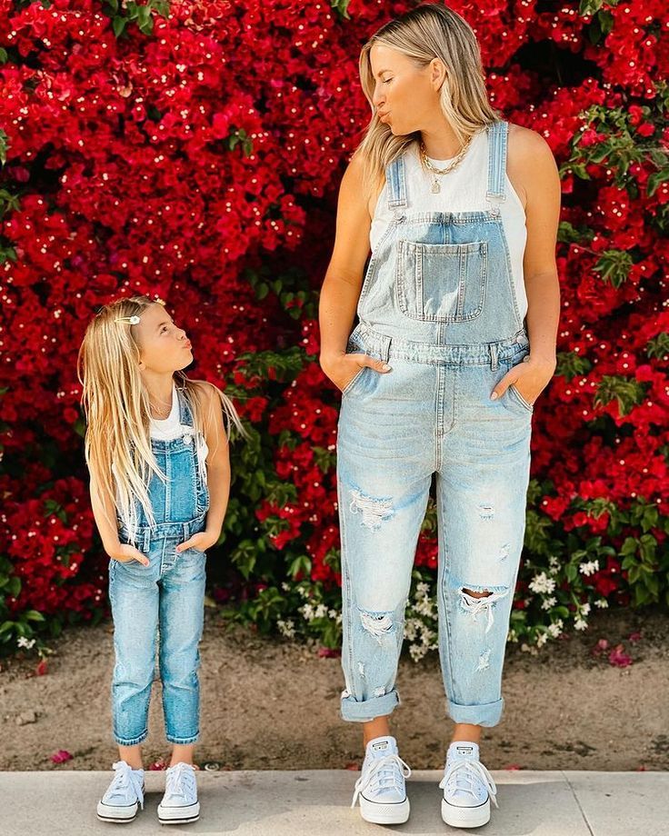 Mother and daughter in matching denim overalls stand in front of vibrant red flowers, sharing a joyful moment.