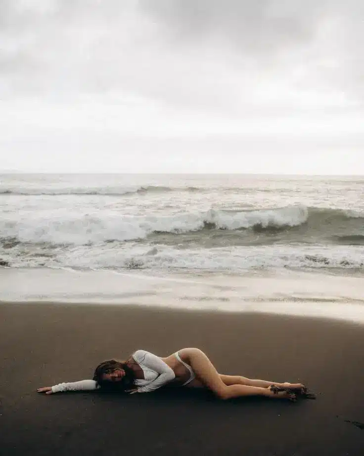 Woman in swimwear lying on a sandy beach near the ocean waves under a cloudy sky, creating a serene coastal scene.