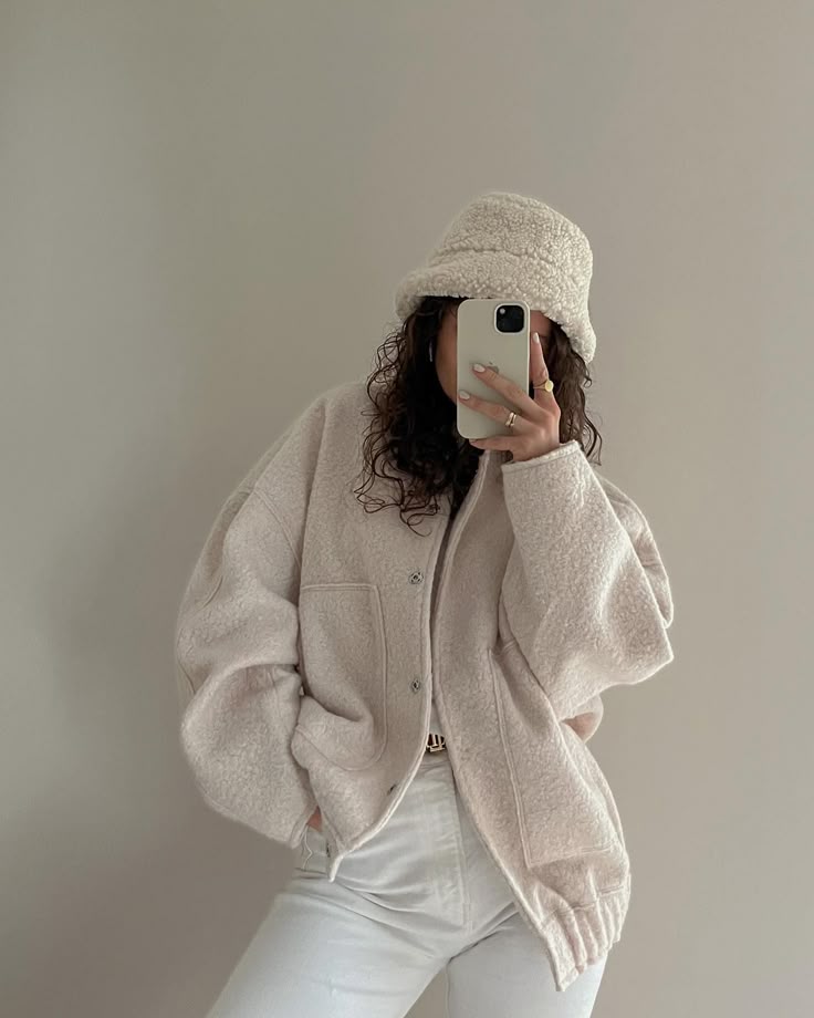 Person in cozy beige jacket and hat taking a mirror selfie with phone, showcasing casual winter fashion style.