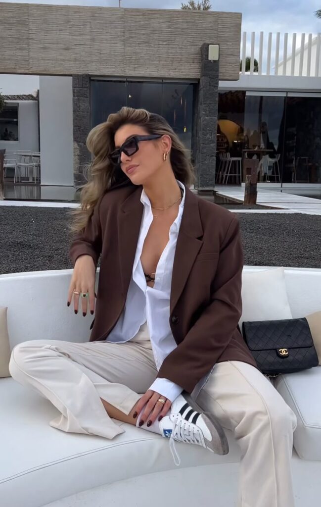 Stylish woman in sunglasses and suit sitting on outdoor sofa with designer handbag, exuding elegance and confidence.