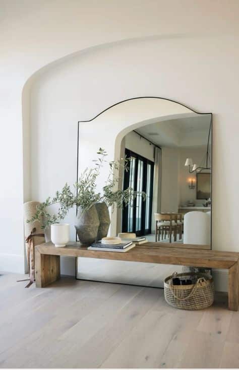 Minimalist entryway with large mirror, wooden bench, decorative plants, and books, creating a serene and stylish ambiance.