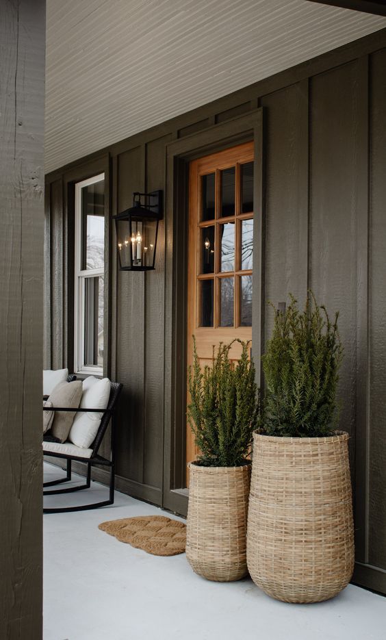 Cozy porch with green plants in wicker baskets, wooden door, and outdoor seating on a white floor.