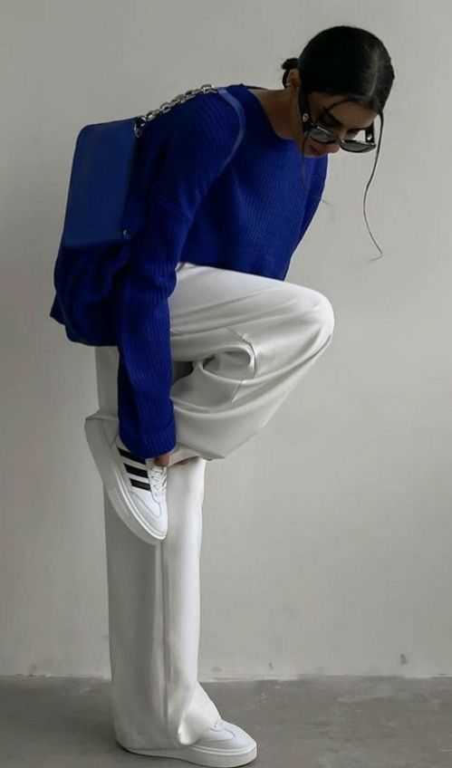 Fashionable person in blue sweater and white pants adjusting sneakers, stylish handbag and sunglasses.