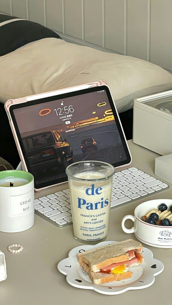 Tablet with a city wallpaper, keyboard, and a breakfast setup including milk, sandwich, and fruit bowl on a desk.