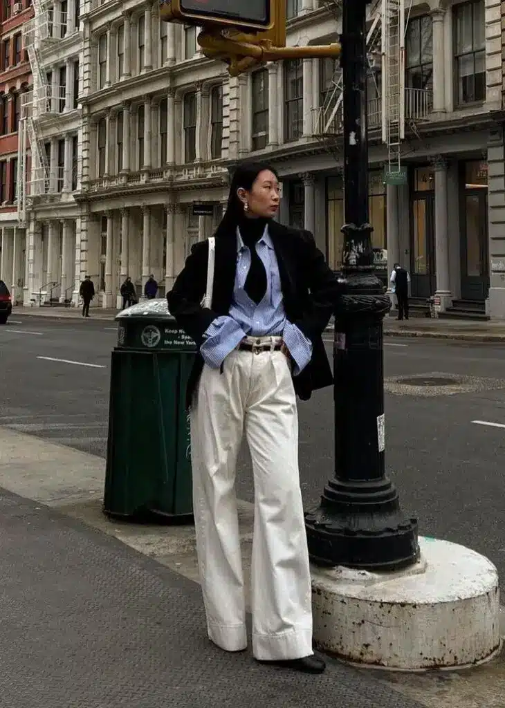 Stylish woman in white trousers and black blazer standing on a city street corner.