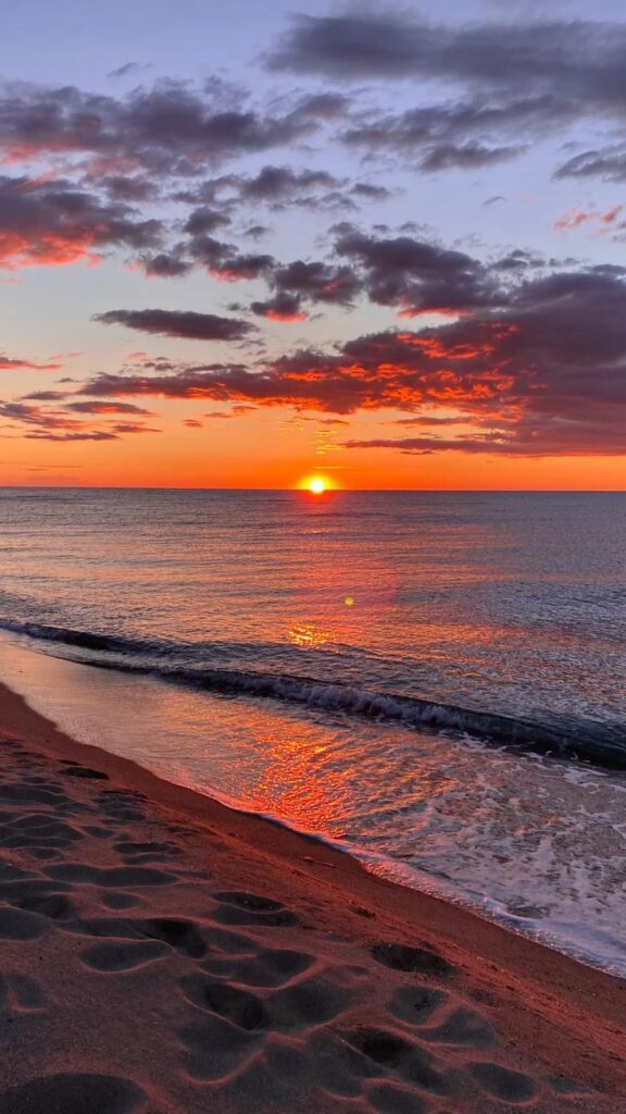 Vibrant beach sunset with orange reflections on water and sandy shore, under dramatic, colorful clouds.