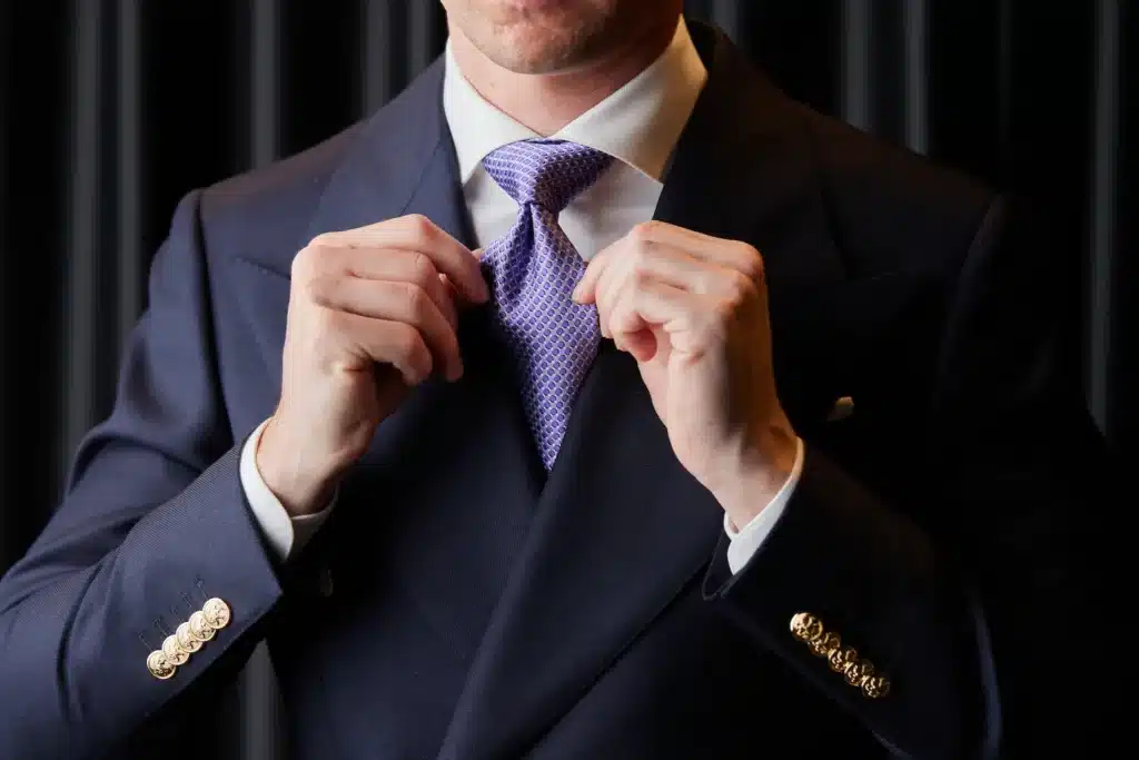 211111_TheBespokeCorner_November-935 Man adjusting purple tie in a dark suit with gold buttons, in front of a curtain background. | Sky Rye Design Man adjusting purple tie in a dark suit with gold buttons, in front of a curtain background.