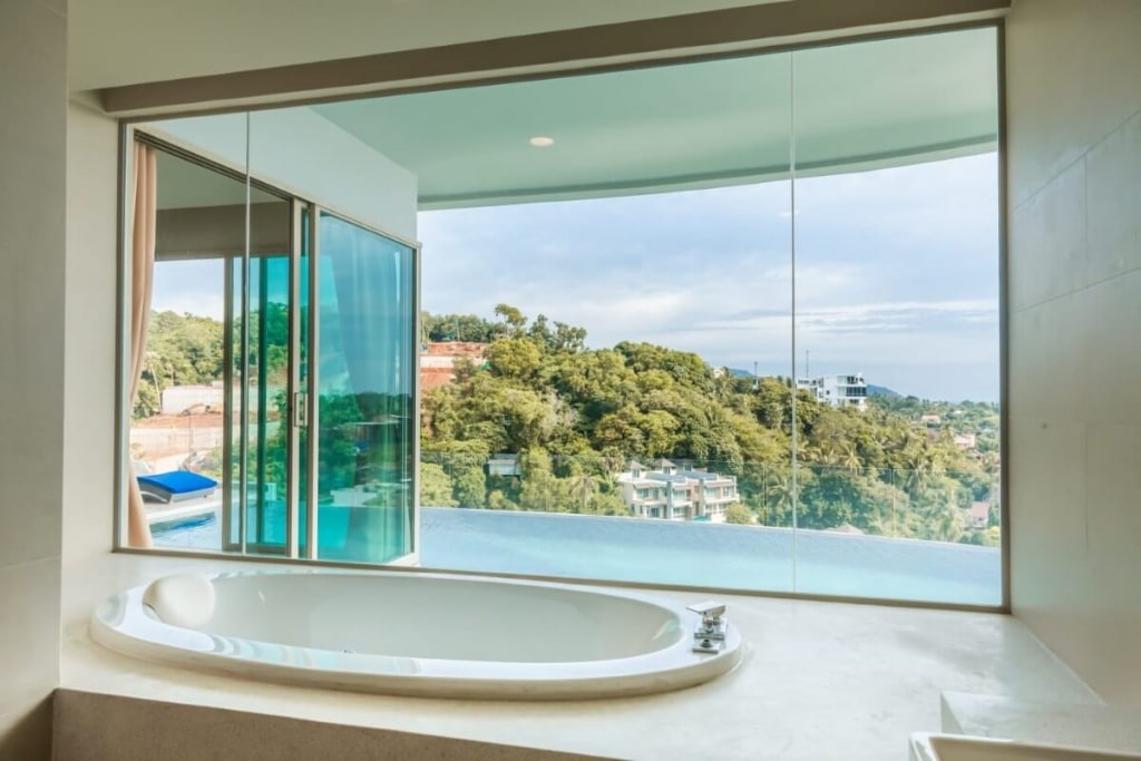Luxurious bathtub with panoramic view of lush greenery and infinity pool through expansive glass window.