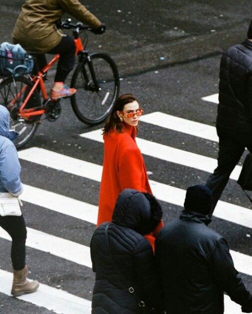 Woman in red coat crossing busy city street with pedestrians and cyclist.