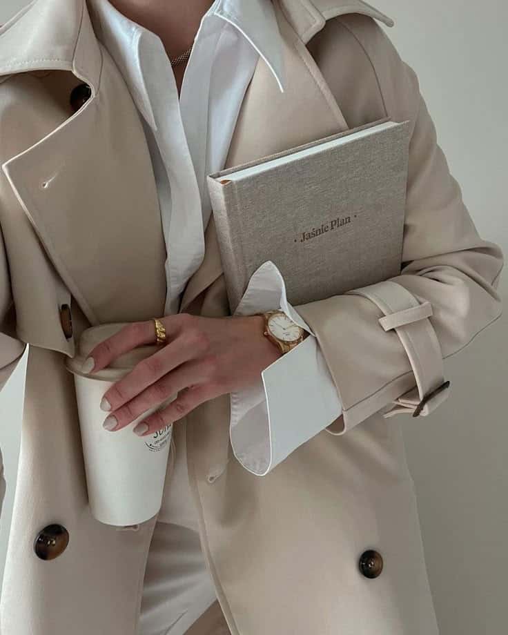 Stylish person in beige trench coat holds a coffee cup and planner, showcasing elegant business attire and accessories.