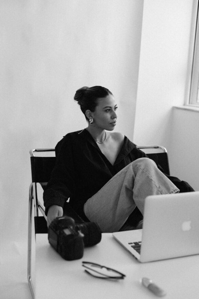 367c1679e9eb4656cc8cb7c86daf5ed4 Woman in black top and jeans sitting at a desk with laptop and camera, looking thoughtfully out the window. | Sky Rye Design Woman in black top and jeans sitting at a desk with laptop and camera, looking thoughtfully out the window.