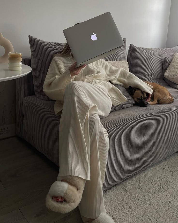 Cozy home office setup: person in white loungewear on sofa with laptop, beside a relaxed dog.
