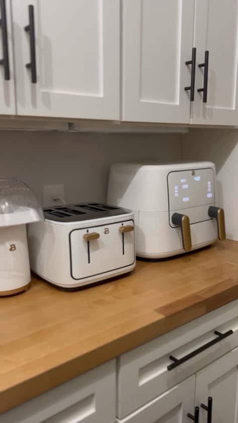 Modern kitchen with white toaster and air fryer on wooden countertop. White cabinets and black handles above.
