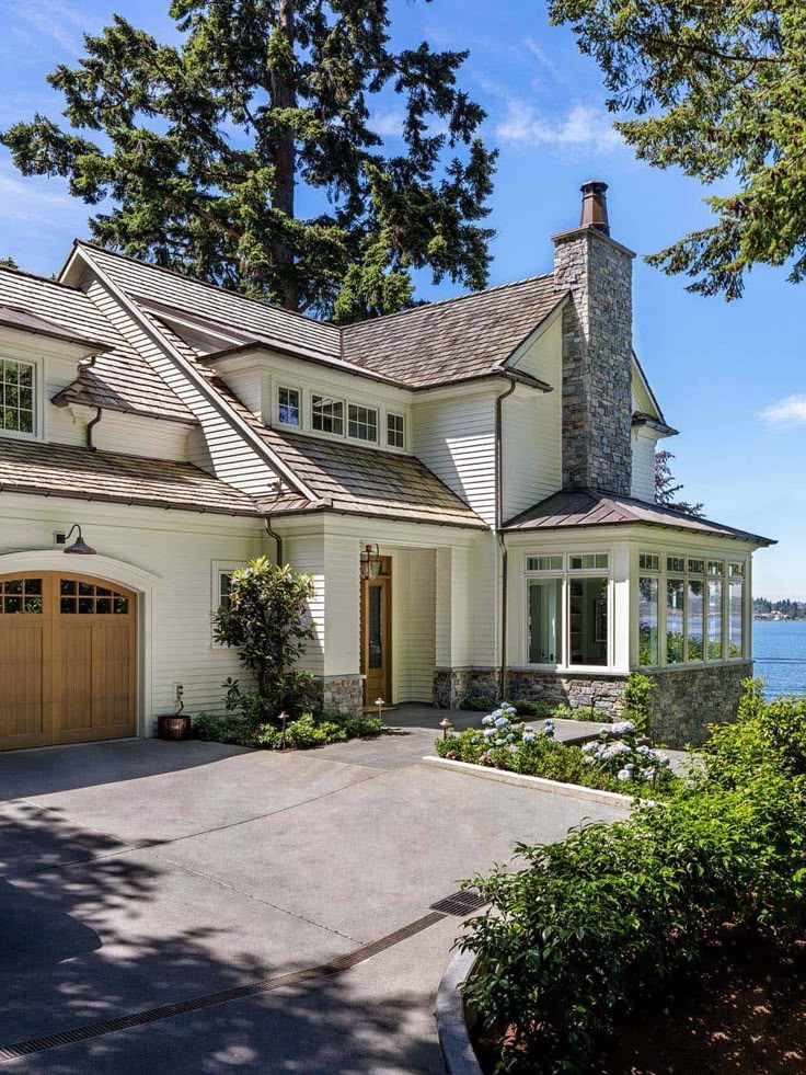 Elegant lakefront house with a stone chimney, large windows, and a wooden garage. Surrounded by tall trees and garden.