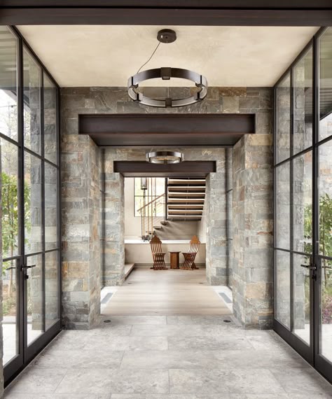 Modern stone hallway with glass doors, leading to a bright room with stairs and wooden chairs. Contemporary interior design.