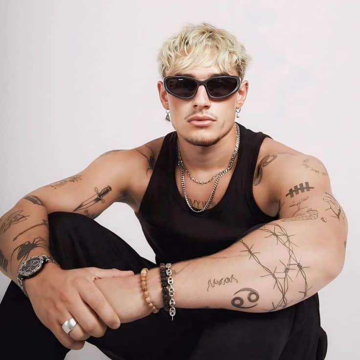 _ 47 Man with blonde hair, sunglasses, and tattoos in black tank top, sitting casually with arms resting on knees. | Sky Rye Design Man with blonde hair, sunglasses, and tattoos in black tank top, sitting casually with arms resting on knees.