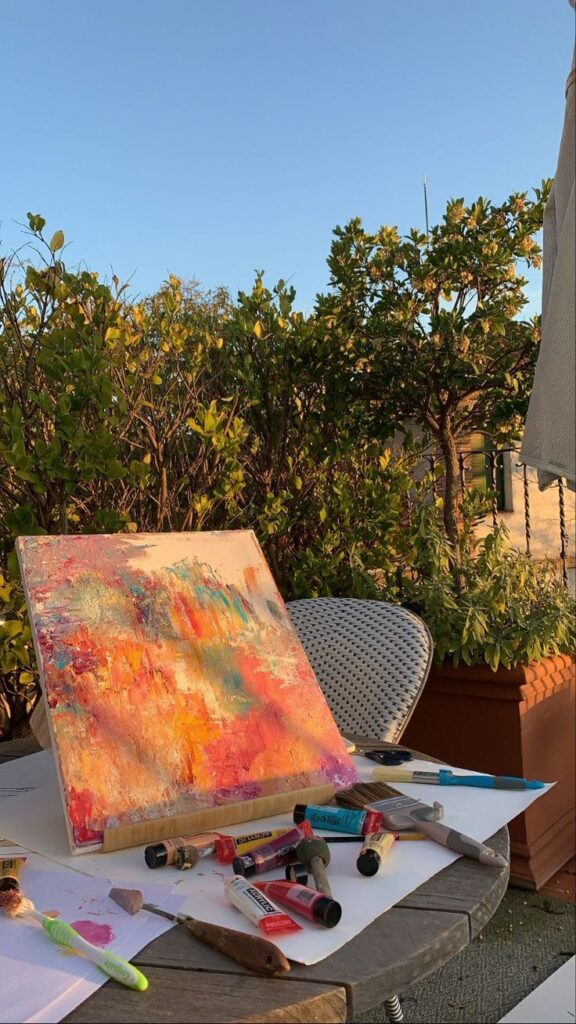 Colorful abstract painting on a table outdoors, surrounded by plants and paint tubes, under a clear blue sky.