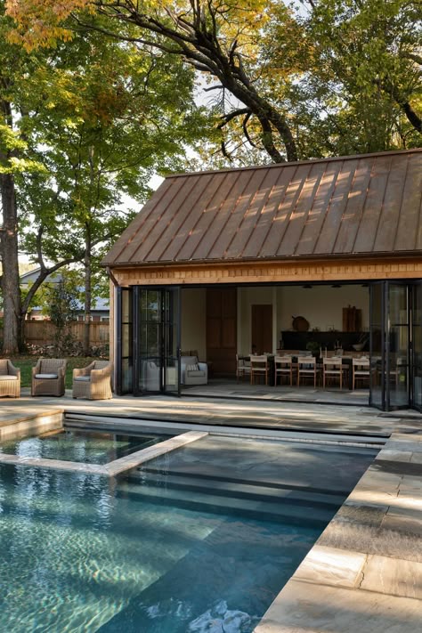 Modern patio with a pool and open-air dining area surrounded by green trees under a clear sky.