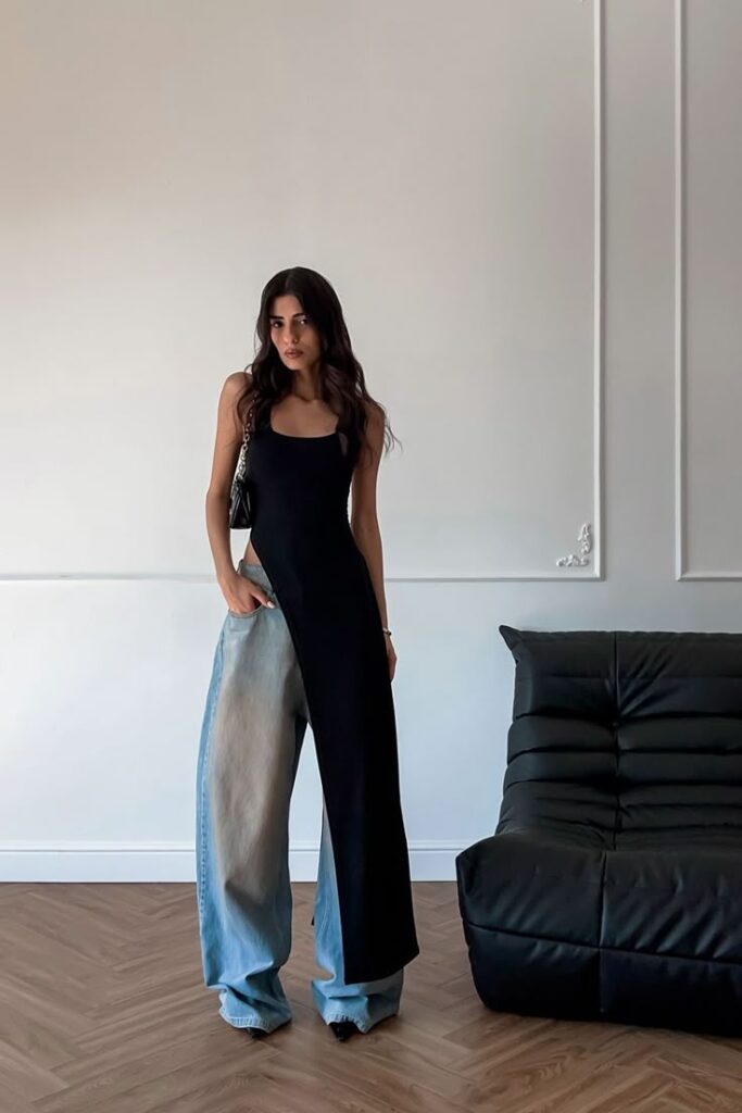 Fashion-forward woman in asymmetrical black dress over wide-leg jeans in modern living room setting.