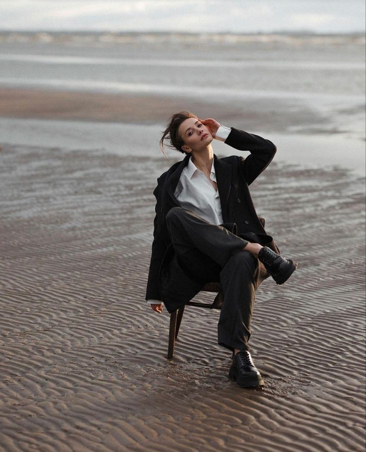 Person in black suit sitting on a chair at the beach, relaxed pose with a serene ocean backdrop.