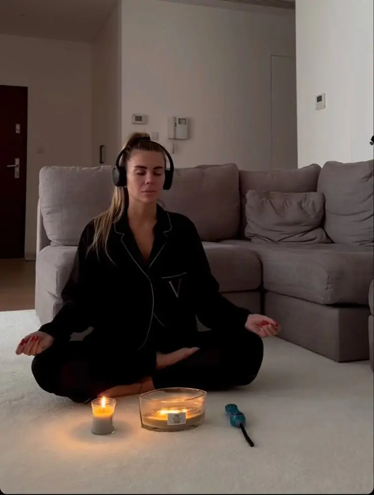 Woman meditating with headphones, sitting on floor near candles in cozy living room.