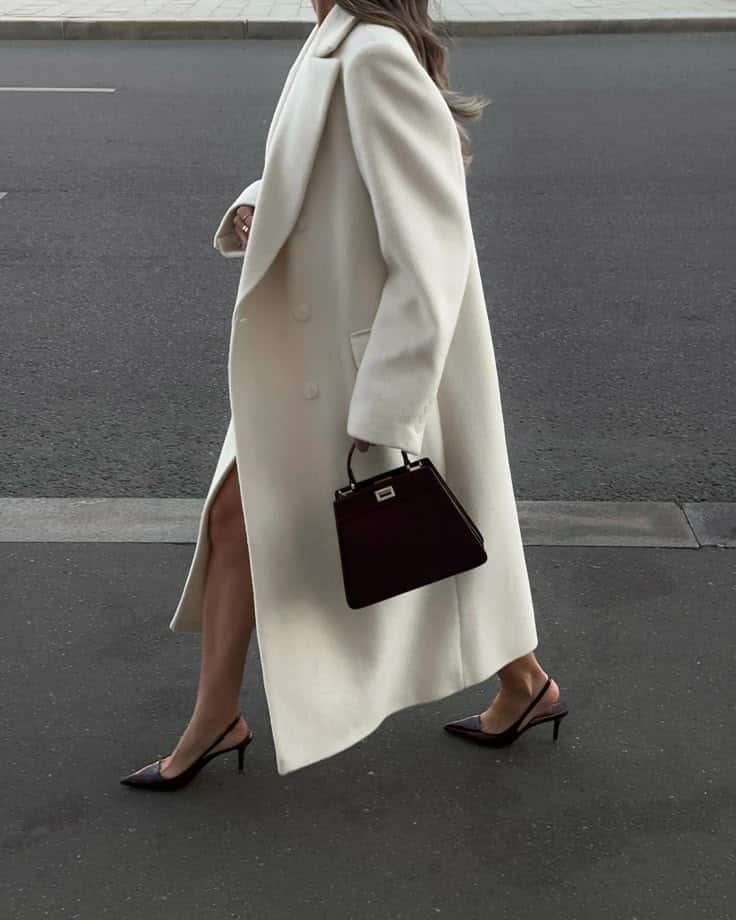 Stylish woman in a long white coat and heels walking on a city street, carrying a chic black handbag.