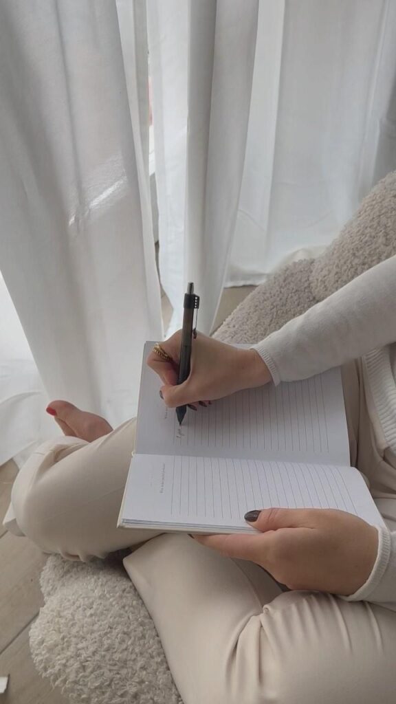 51b69a69-5144-463c-8a56-f4725a5b343b Person writing in a notebook on a cozy chair near a window with sheer curtains, capturing a peaceful moment. | Sky Rye Design Person writing in a notebook on a cozy chair near a window with sheer curtains, capturing a peaceful moment.