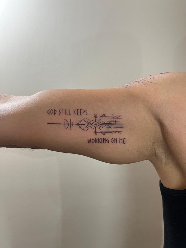 Arm with intricate tattoo design and the text God still keeps working on me on bicep against a neutral background.