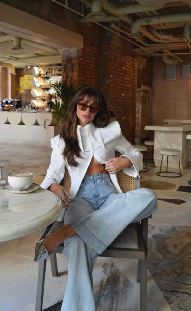 Woman in a stylish white blazer and jeans sitting in a modern cafe, wearing sunglasses and holding a coffee cup.