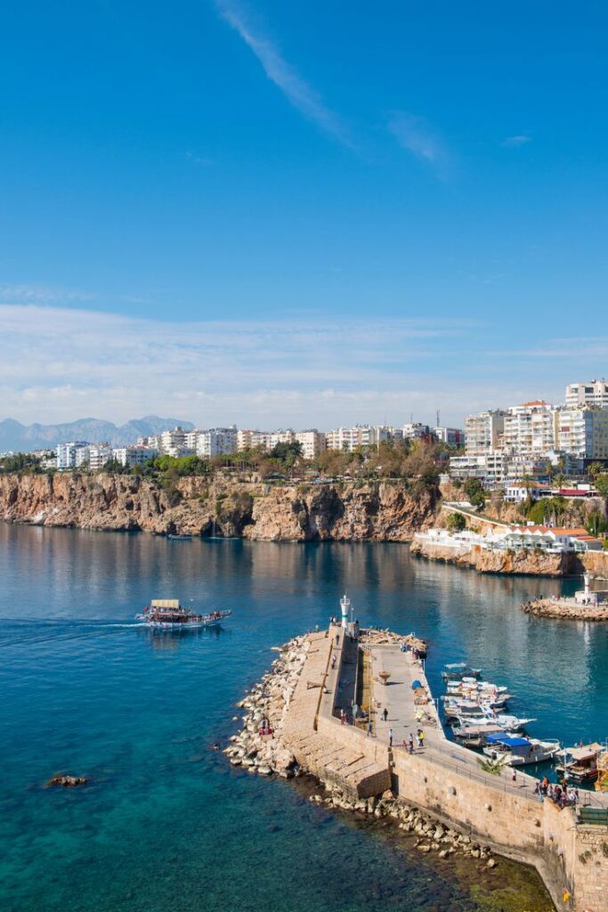 Scenic coastal view of vibrant harbor with boats and lighthouse, surrounded by cliffs and urban skyline under clear blue sky.