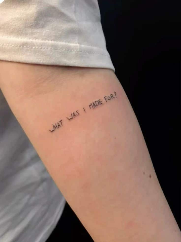 Minimalistic arm tattoo with the text WHAT WAS I MADE FOR? in fine line style, reflecting introspective thought.