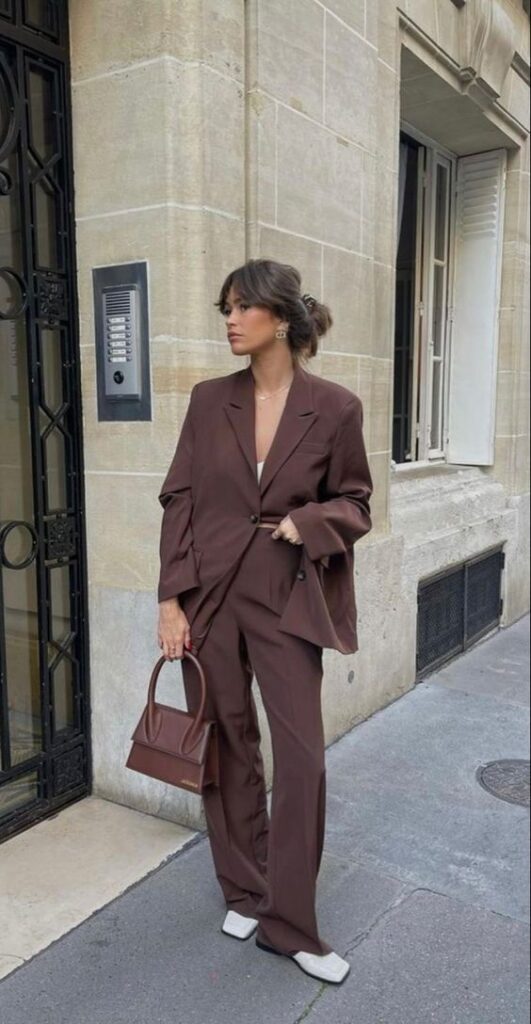 Woman in a stylish brown suit stands outside, holding a matching handbag. Chic urban fashion vibe.