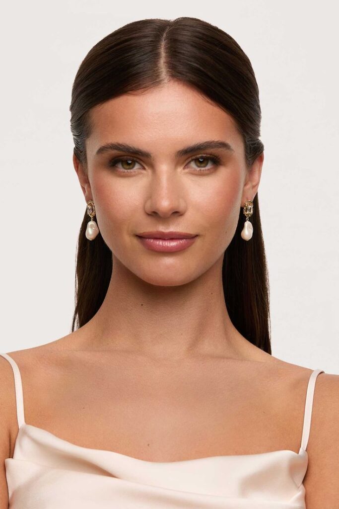 Woman with pearl earrings and smooth brown hair wearing a white top, showcasing elegant fashion style.