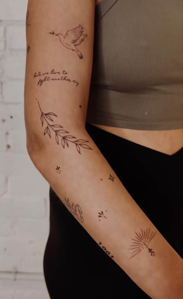 Arm with minimalist tattoos: bird, text, leaves, and sunburst. Casual style with a tank top as background.