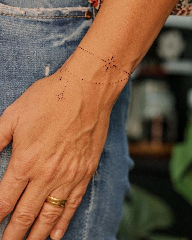 Minimalist compass star wrist tattoo on a hand with a gold ring, wearing casual denim jeans in a relaxed setting.