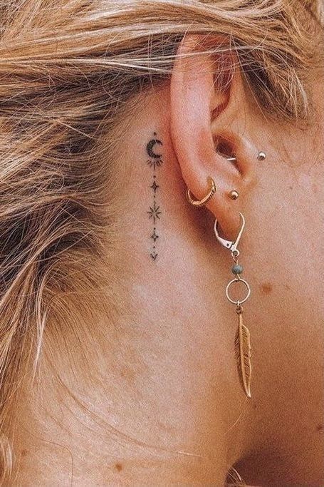 Ear close-up with celestial tattoo, crescent moon, stars; multiple earrings including hoops and feather pendant. Bohemian style.