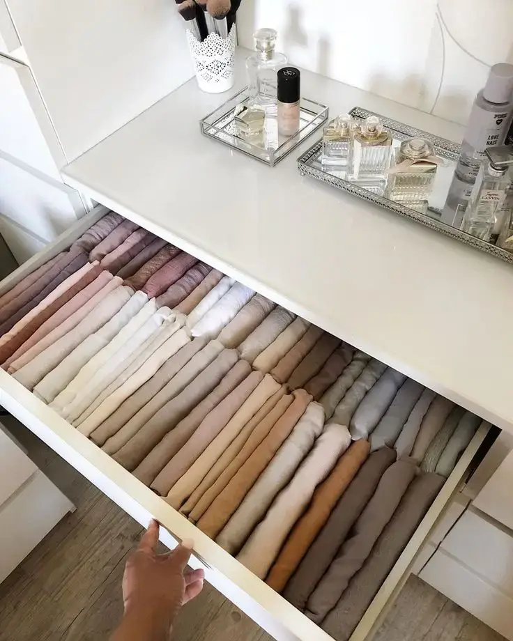 Organized drawer with neatly folded clothes in soft colors, makeup brushes, perfume, vanity essentials on top.