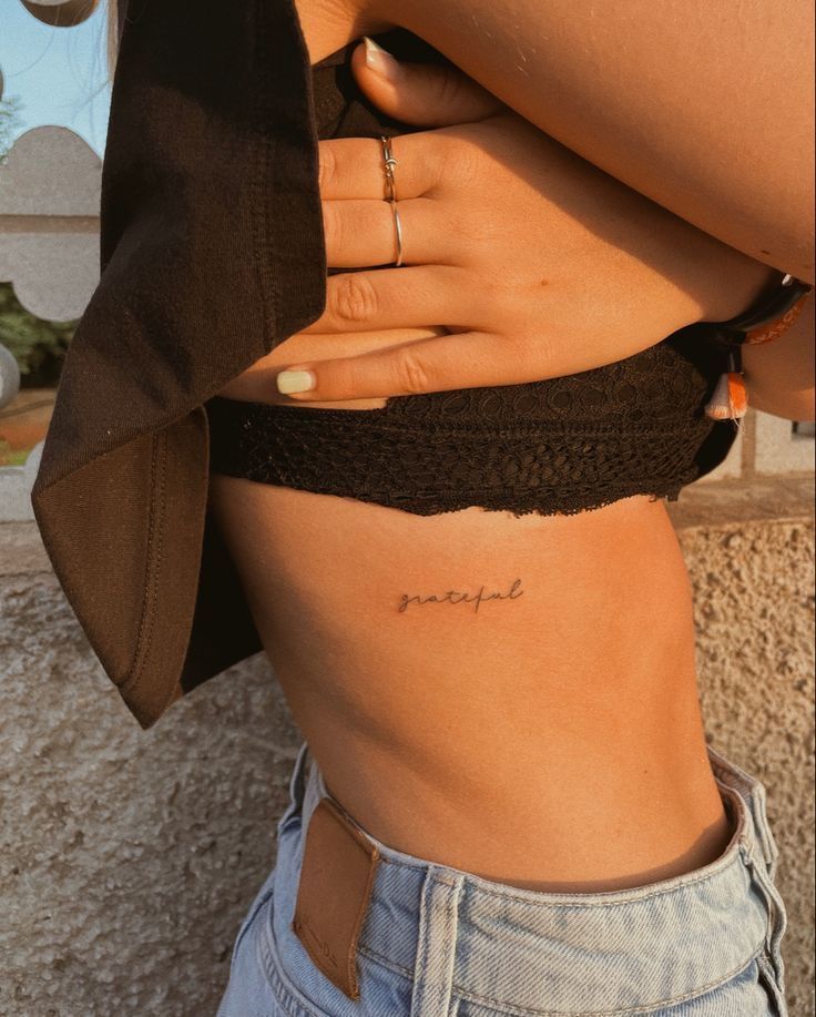 Woman with grateful tattoo on ribcage, wearing denim jeans and black top, showcasing minimal tattoo style.