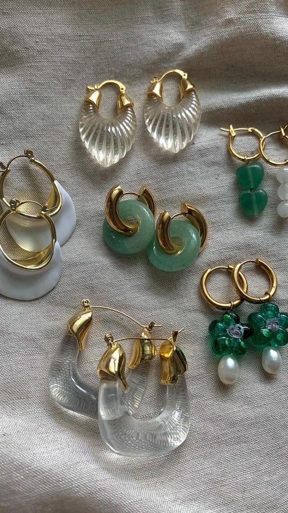Assorted stylish gold and crystal earrings on a fabric background, featuring green and pearl accents.