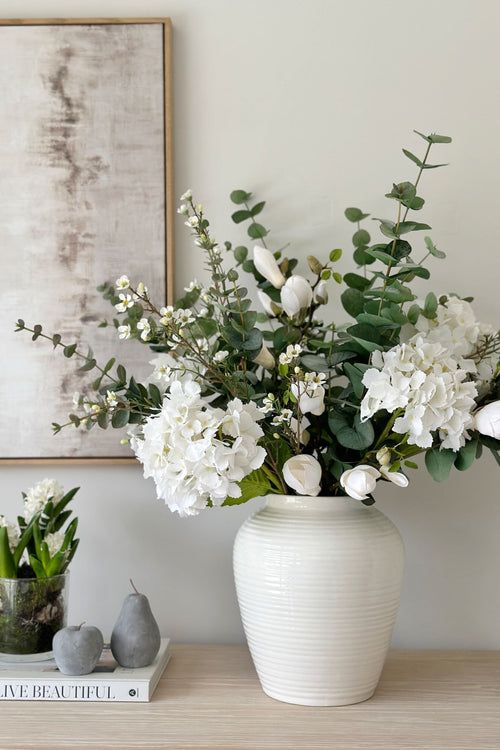 Elegant white vase with lush floral arrangement on a wooden table against a soft-colored wall.
